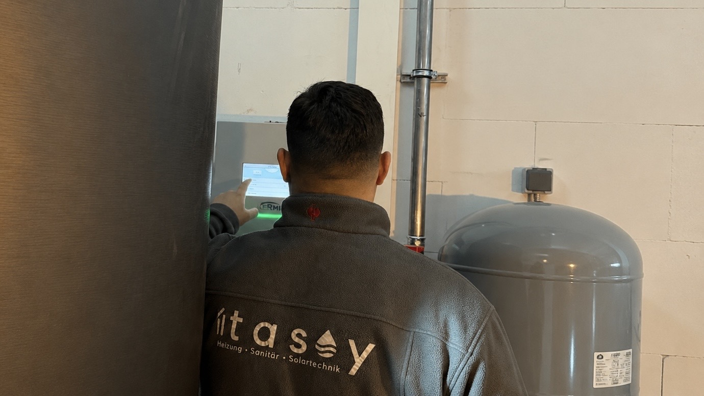 Atasoy Monteur im Firmen-Fleece bedient die Regelung einer Heizungsanlage im Technikraum